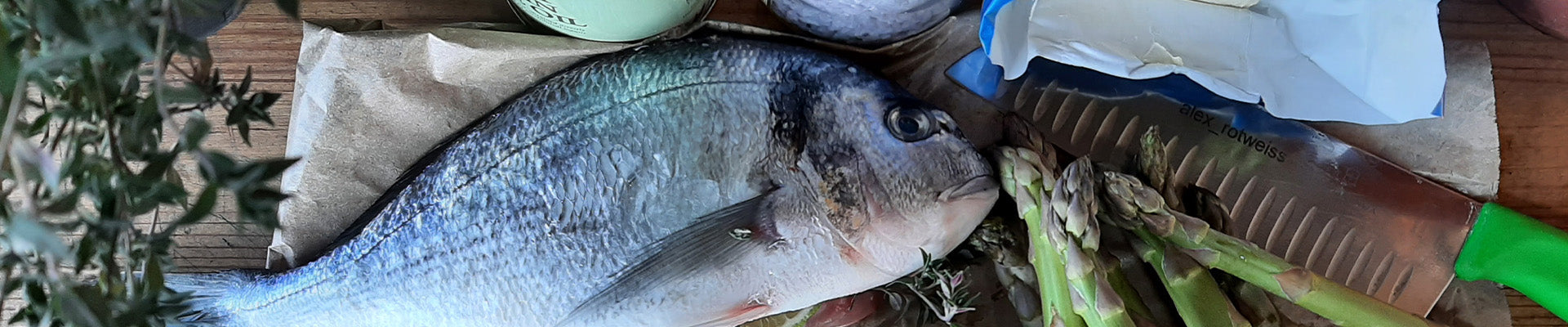 Zu sehen sind eine frische Dorade, ein Messer und die Spitzen von grünem Spargel. A fresh Dorado, a knife and the tips of green asparagus can be seen.